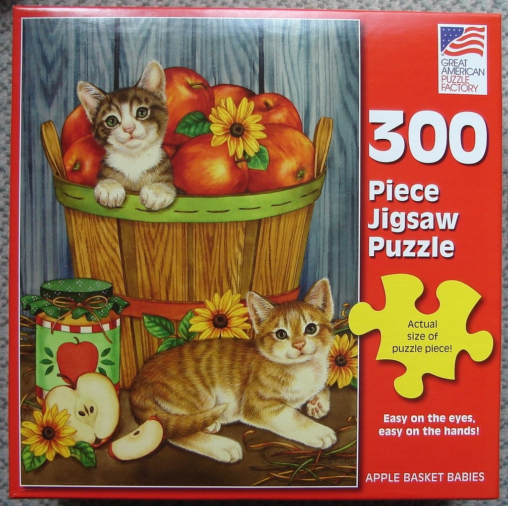 Two kittens, one sitting in a bushel basket of apples and one resting on the ground next to a jar of apple preserves.