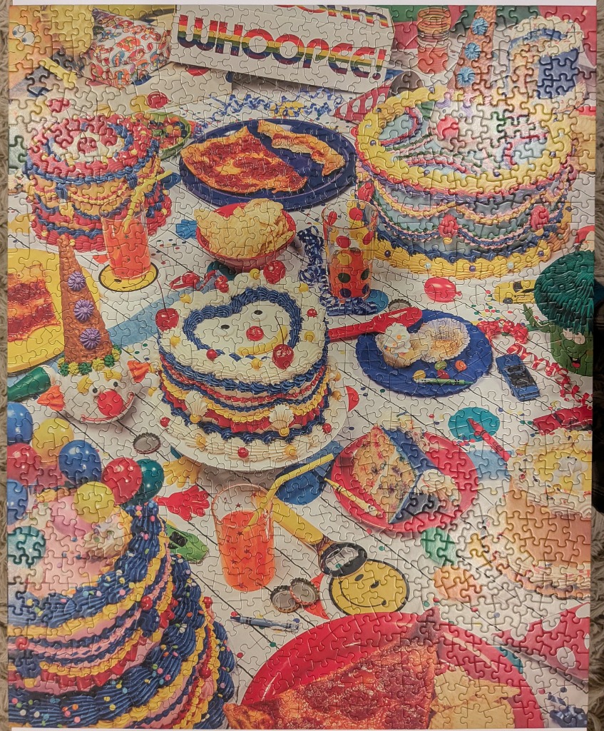 An overhead view of a chaotic, brightly colored birthday party table filled with cakes, pizza, and party snacks.