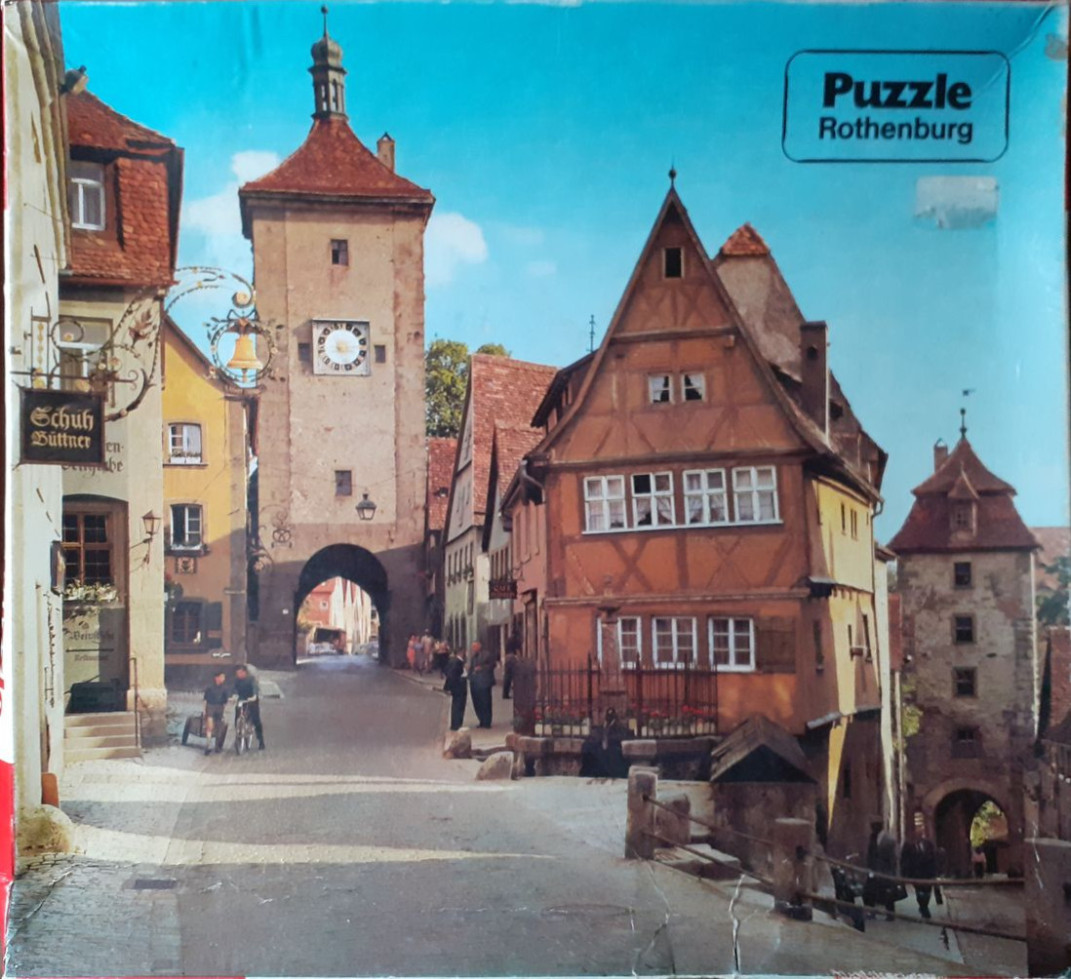 A view of the medieval town of Rothenburg with timber-framed houses, a clock tower, and a stone archway under a blue sky.