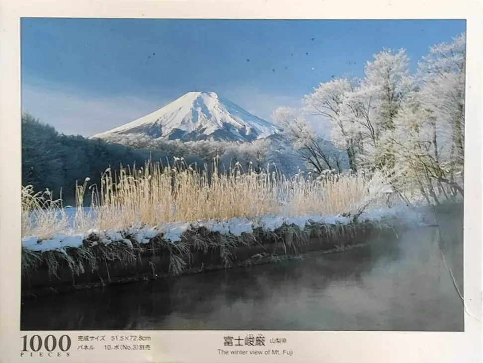 A scenic photograph of a snow-capped Mount Fuji rising above a frozen, mist-covered riverbank in winter.