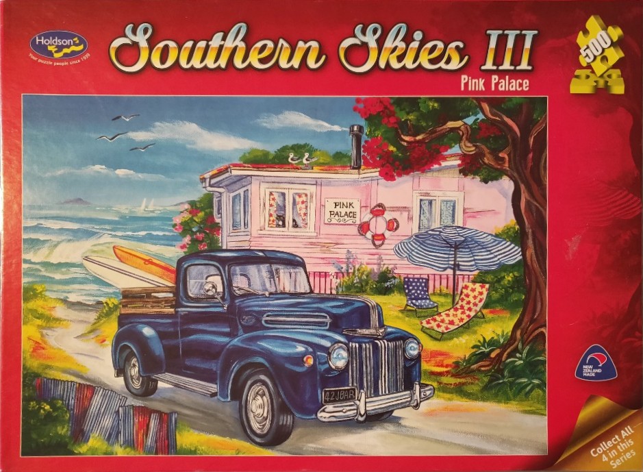 A vintage blue pickup truck parked in front of a coastal pink caravan titled Pink Palace by the ocean.