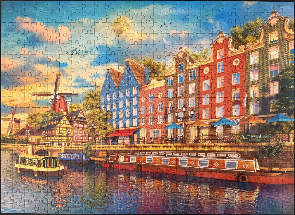 A picturesque European canal scene featuring colorful narrow townhouses, a traditional windmill, and boats at sunset.