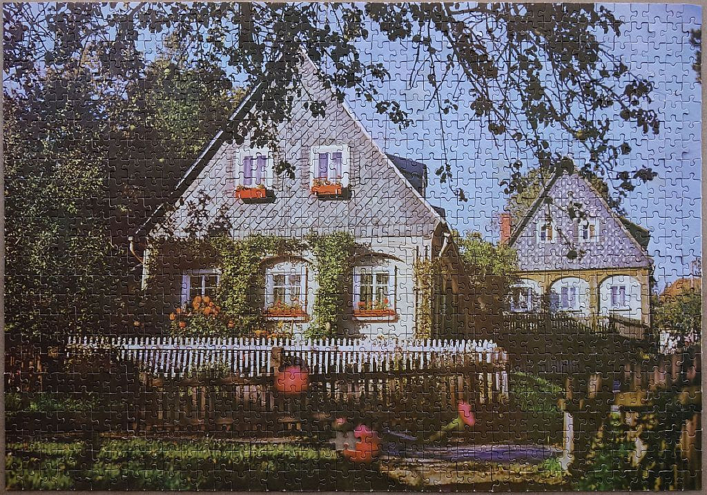 A charming house with slate-tiled gables surrounded by lush greenery and a white picket fence.