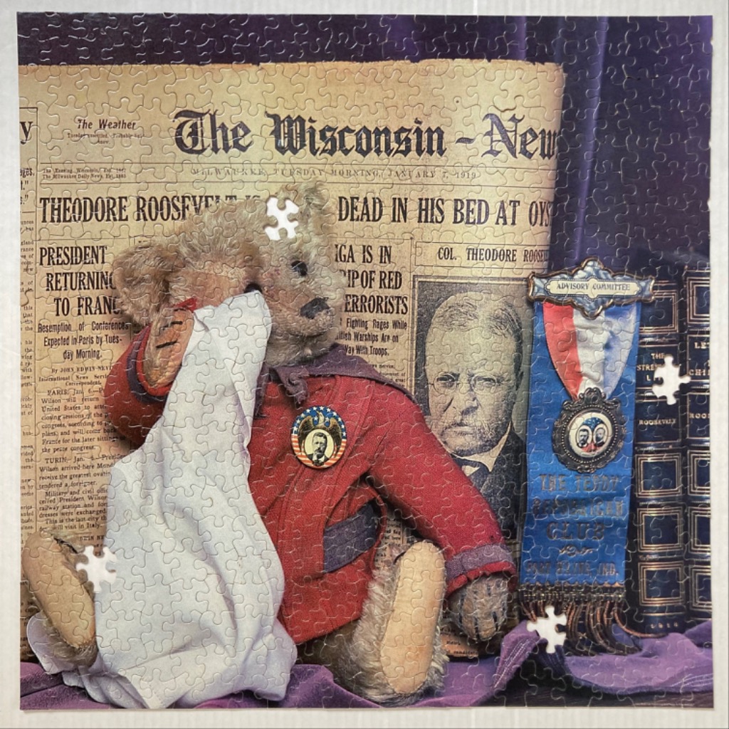 A vintage-style teddy bear wearing a red jacket sits in front of a newspaper front page announcing Theodore Roosevelt