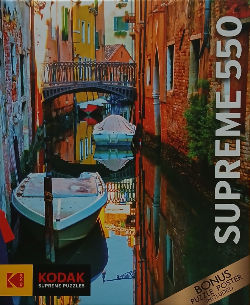 A narrow Venice canal scene with two small boats and a pedestrian bridge between weathered brick and yellow buildings.
