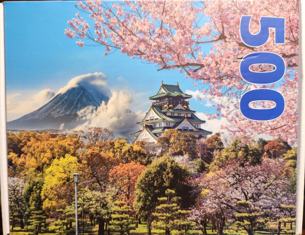A Japanese castle surrounded by cherry blossoms with Mount Fuji in the background under a blue sky.