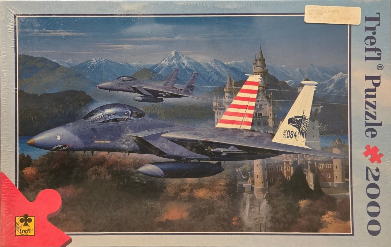 Two F-15 fighter jets flying over a European-style castle set against a mountain range.