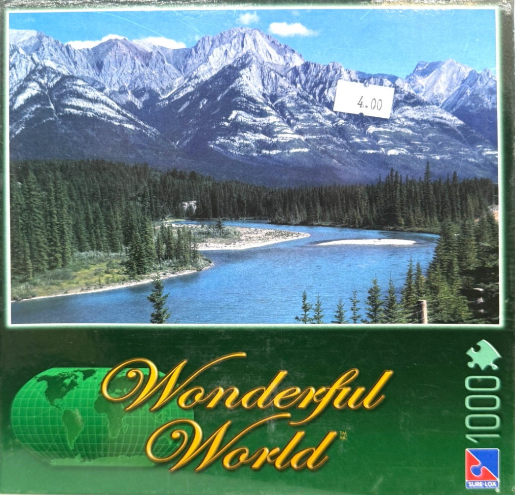 A panoramic landscape photo of a blue river flowing before a massive, snow-dusted mountain range in a coniferous forest.