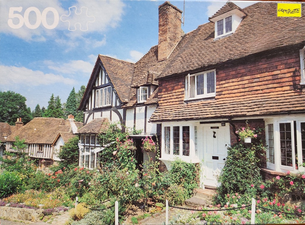 A charming Tudor-style cottage with a red-tiled roof and vibrant flower gardens on a sunny day.