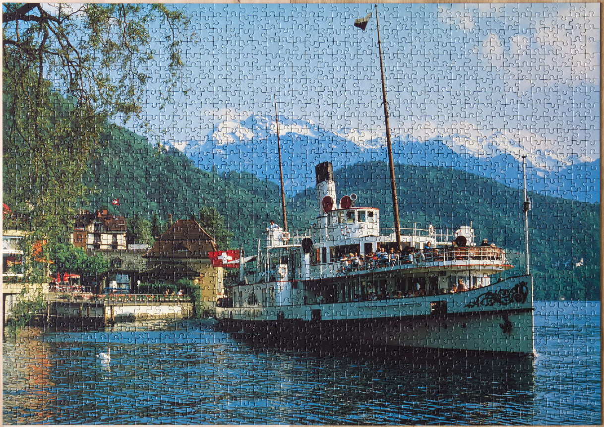 A vintage white paddle steamer boat sailing on a calm lake in front of alpine mountains.