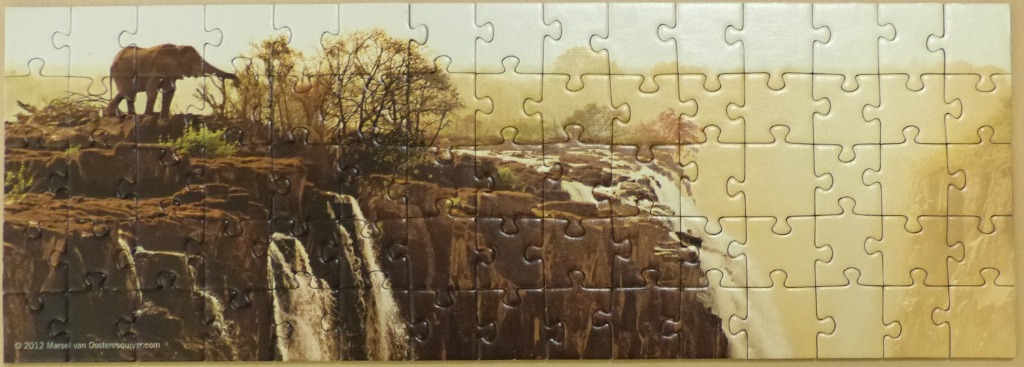 A lone elephant standing on a rocky ridge overlooking a massive waterfall in a golden-hued African landscape.
