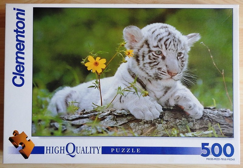 A close-up photograph of a white tiger cub resting on a fallen log in a grassy field with yellow wildflowers.