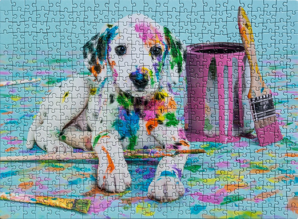 A cute puppy covered in colorful paint splatters sits next to a pink paint can and a paintbrush.
