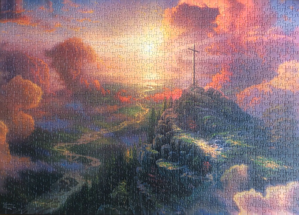 A lone wooden cross atop a grassy hill overlooking a winding river valley during a brilliant sunset.