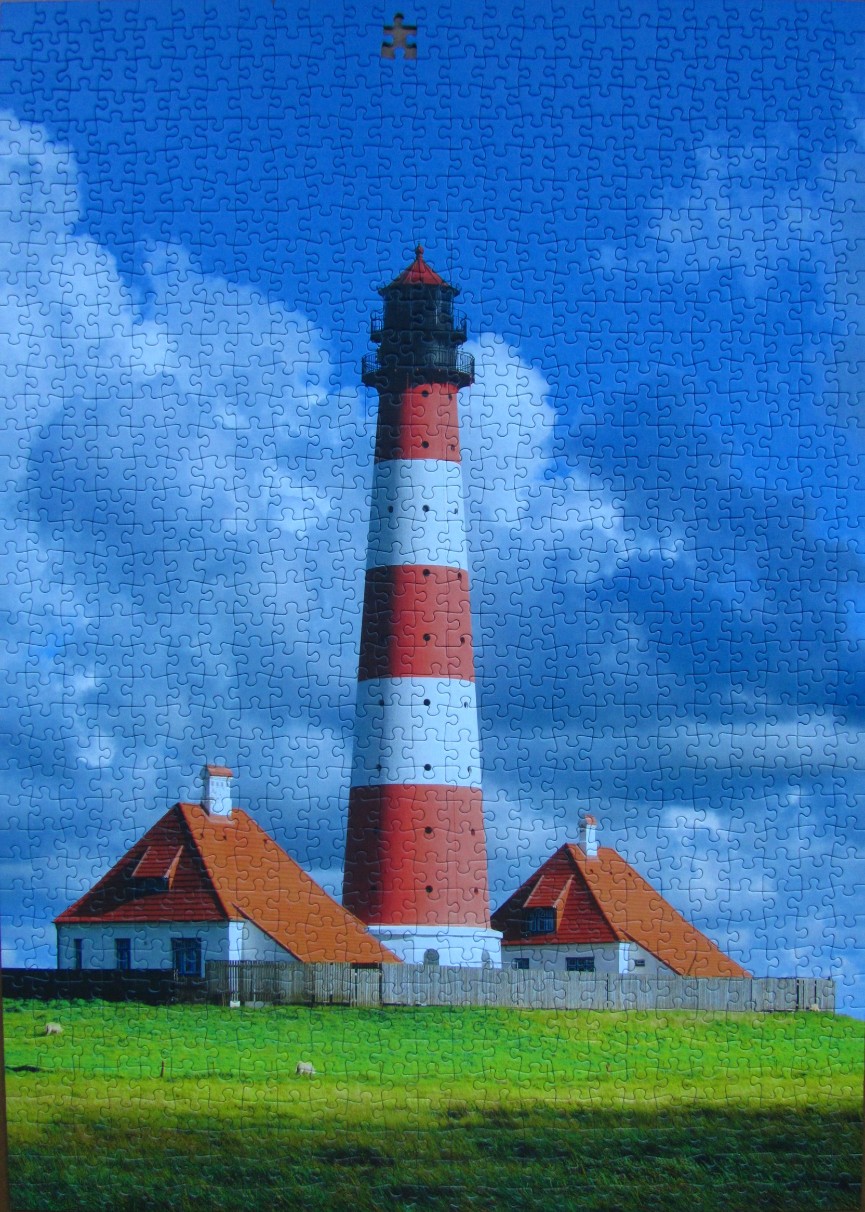 A tall red and white lighthouse stands behind two small houses against a deep blue sky with fluffy white clouds.