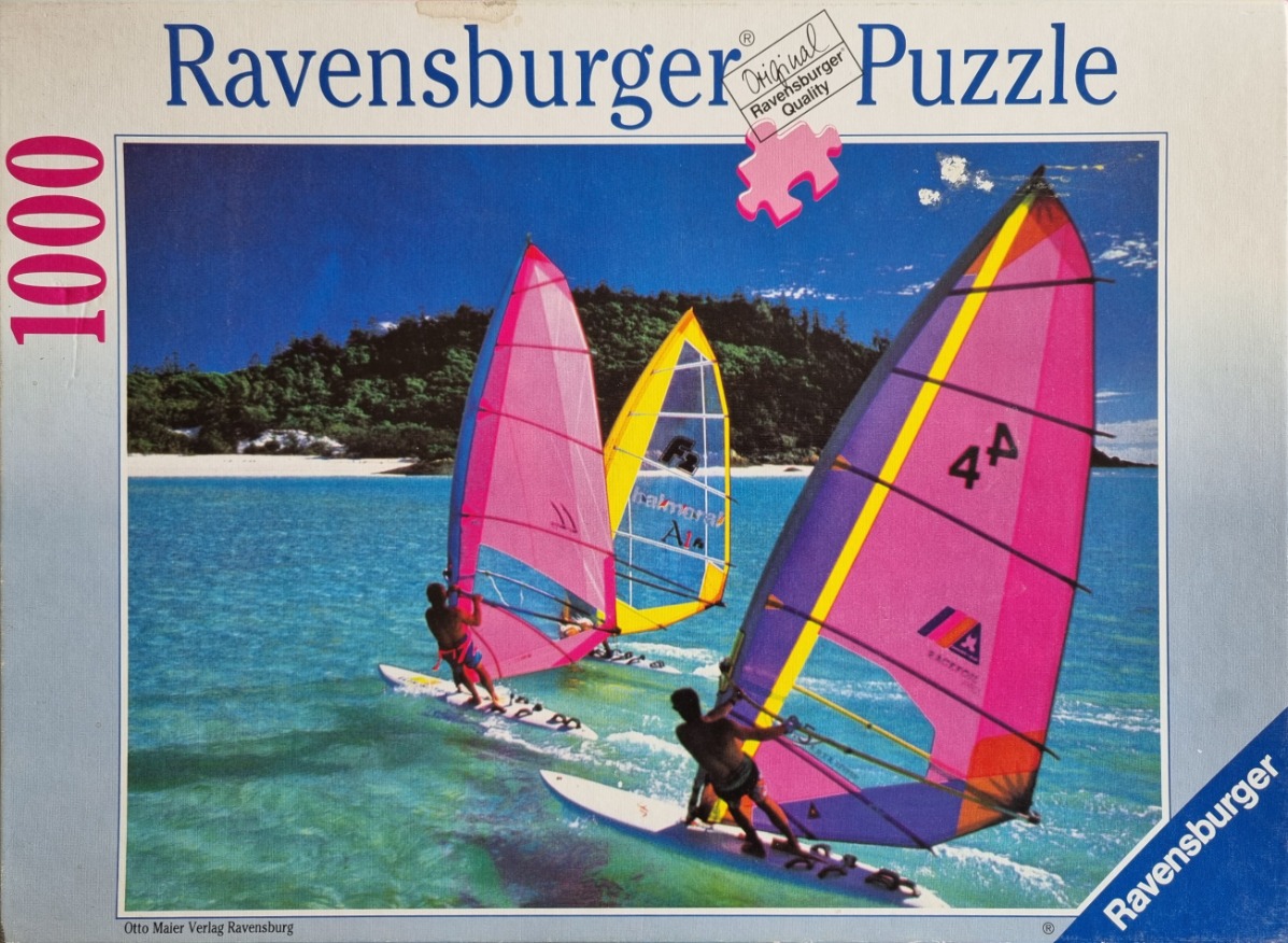 Three windsurfers with colorful sails on bright turquoise water near a white sand beach.