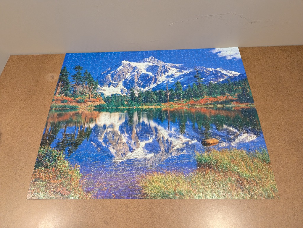 A scenic landscape photo of a snow-capped mountain reflecting in a calm lake surrounded by trees and grasses.