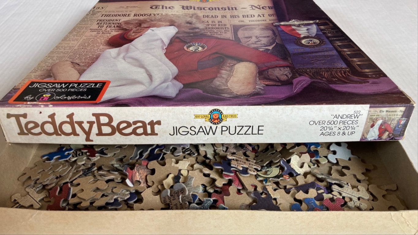 A teddy bear wearing a red jacket and holding a white cloth sits in front of a vintage newspaper and a blue banner.