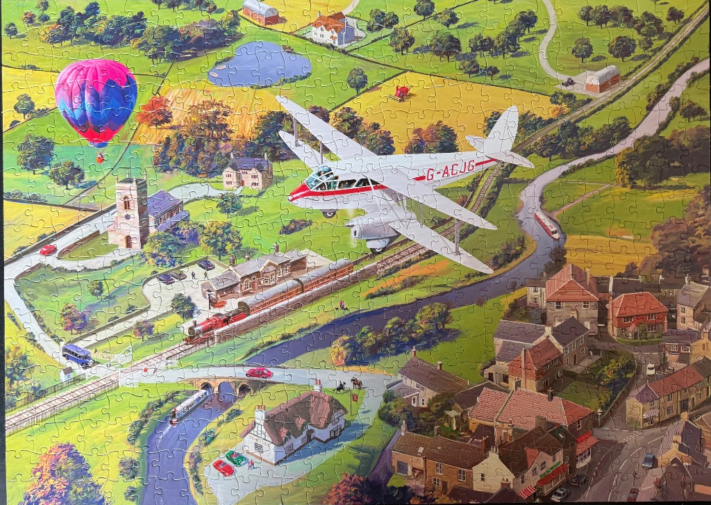 A vintage biplane flies over a nostalgic English countryside scene with a village, railway, canal, and hot air balloon.