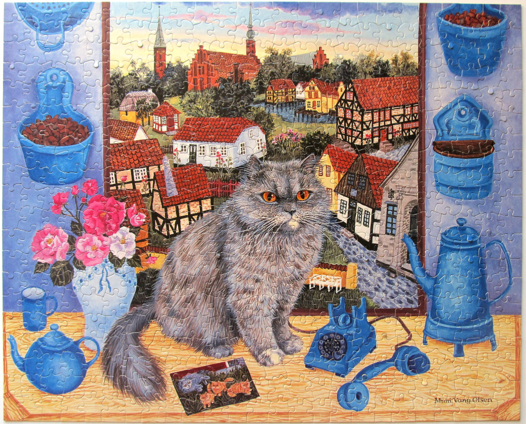 A fluffy grey cat sitting on a table in front of a window overlooking a European village with red-roofed houses.