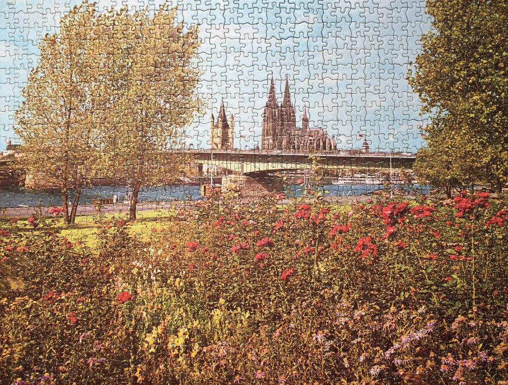 A riverside view of the Cologne Cathedral framed by trees and a foreground garden of red and yellow flowers.