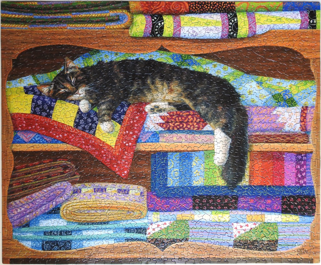 A long-haired cat sleeping on a wooden shelf among stacks of colourful patterned fabric and quilts.