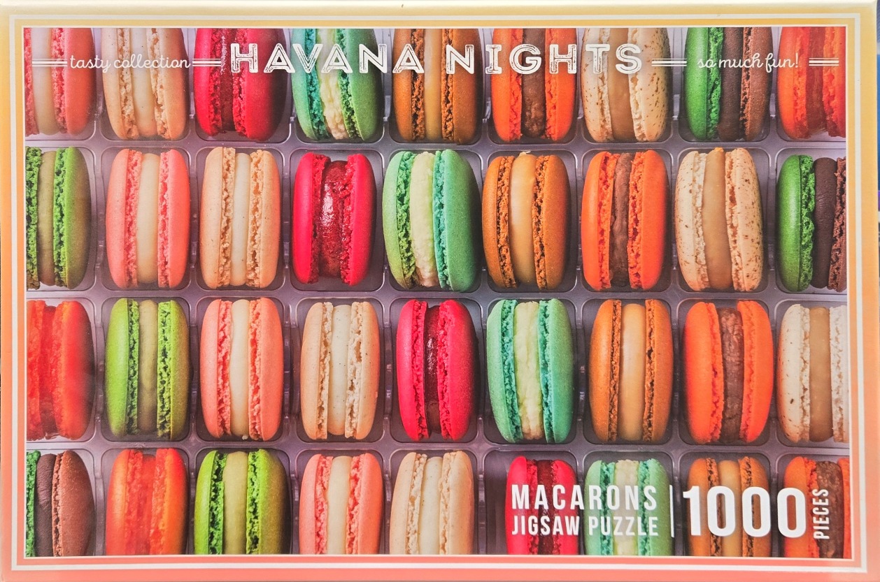 A close-up photograph of a grid of colorful French macarons neatly arranged in a plastic tray.