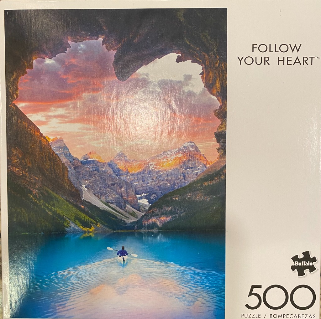 A lone kayaker on a blue lake looking through a heart-shaped cave opening at jagged mountains at sunset.