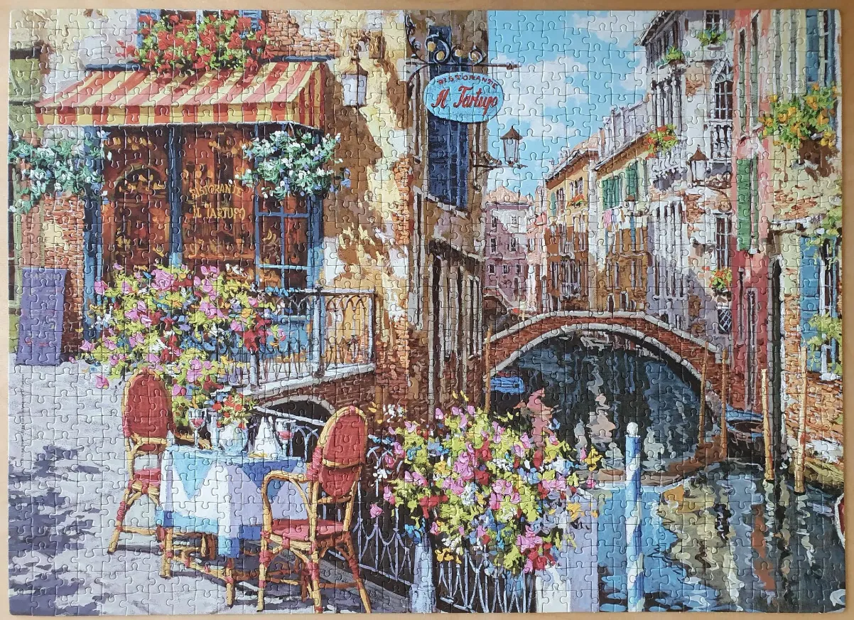 A charming outdoor cafe scene by a canal in Venice, featuring tables, flowers, and old stone buildings.