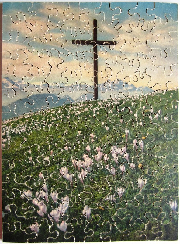 A simple wooden cross standing on a grassy hill covered in white crocuses with mountains in the background.