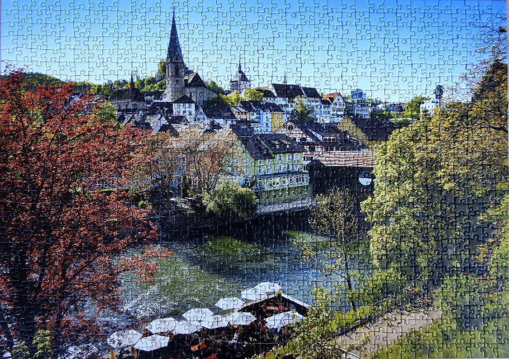 A charming Swiss-style town nestled by a river with a church spire and a cafe with white umbrellas.