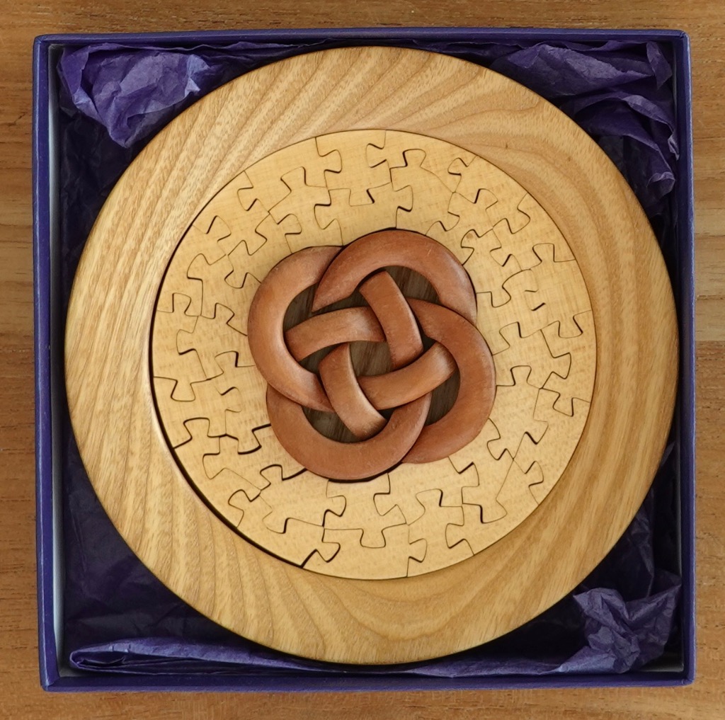 A wooden circular Celtic knot design mounted on a round wooden base inside a purple-lined box.