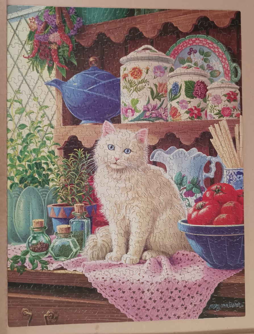 A fluffy white cat sits on a counter surrounded by kitchen herbs, flowers, and ceramic canisters.