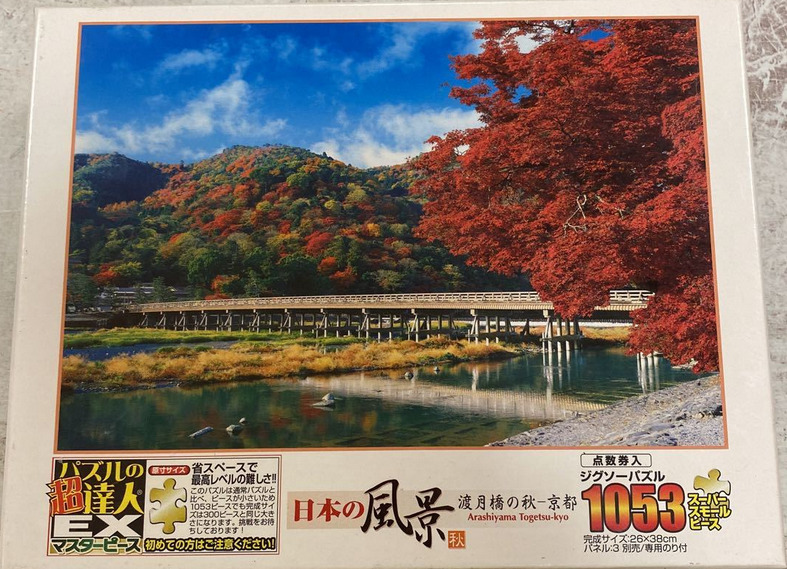 Photographic view of the Togetsukyo Bridge in Kyoto surrounded by vibrant autumn foliage under a blue sky.