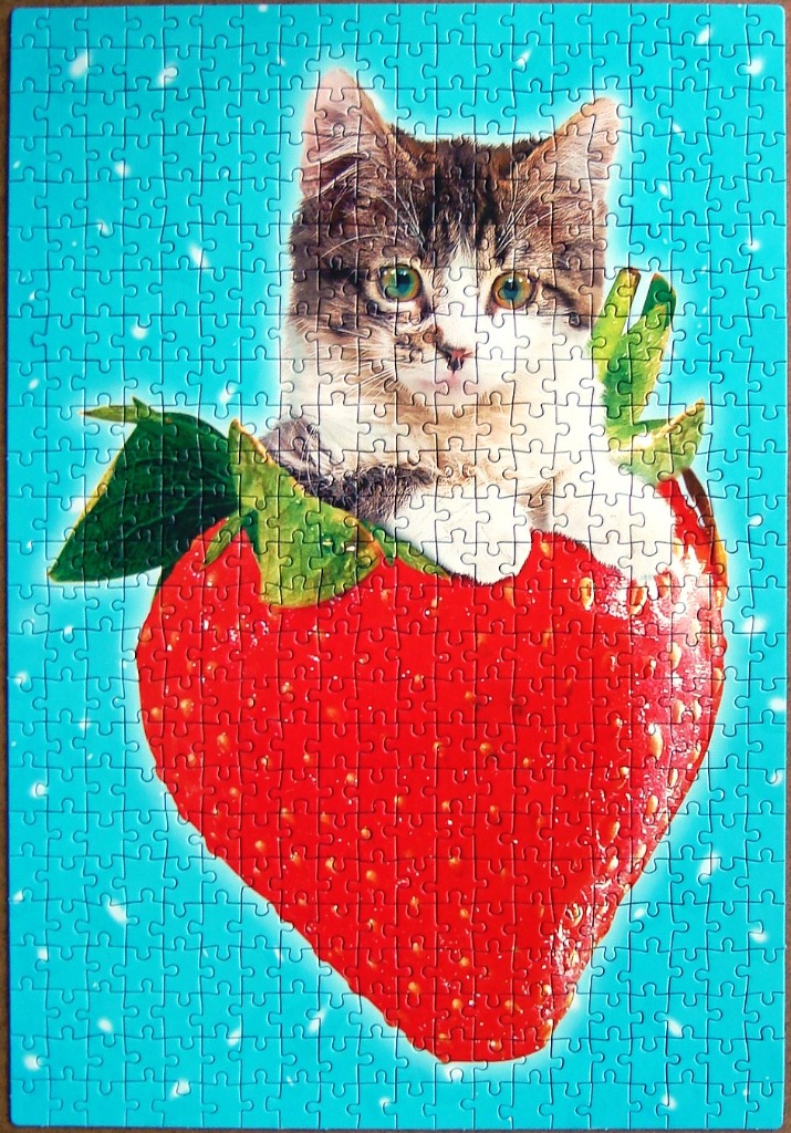 A cute tabby kitten peaking out from inside a large, ripe red strawberry against a bright blue background.