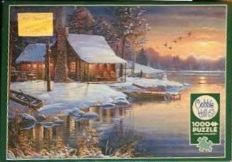 A snow-covered log cabin at sunset beside a calm lake with geese flying in the distance.