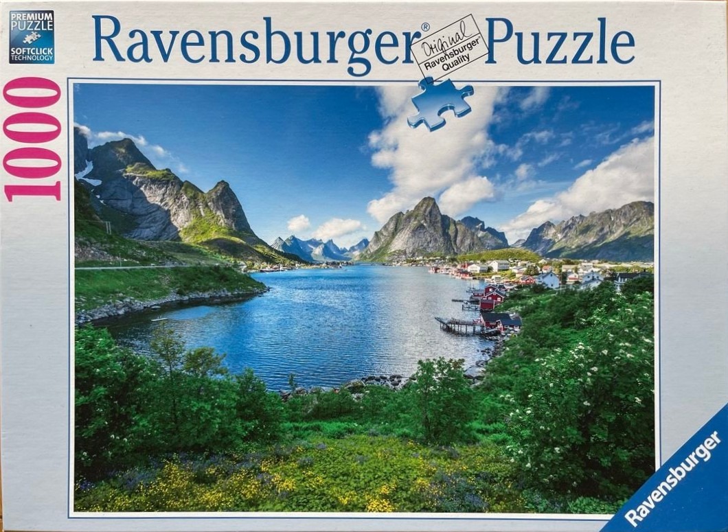 A panoramic landscape photo of Reine, Norway, featuring a deep blue bay surrounded by jagged mountains and red houses.