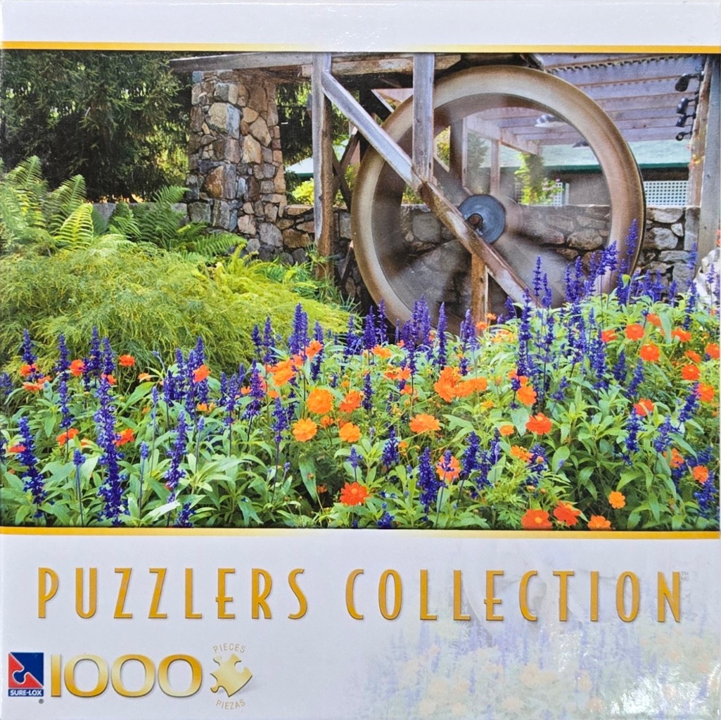 A large rusted water wheel behind a vibrant garden bed of purple salvia and orange cosmos flowers.