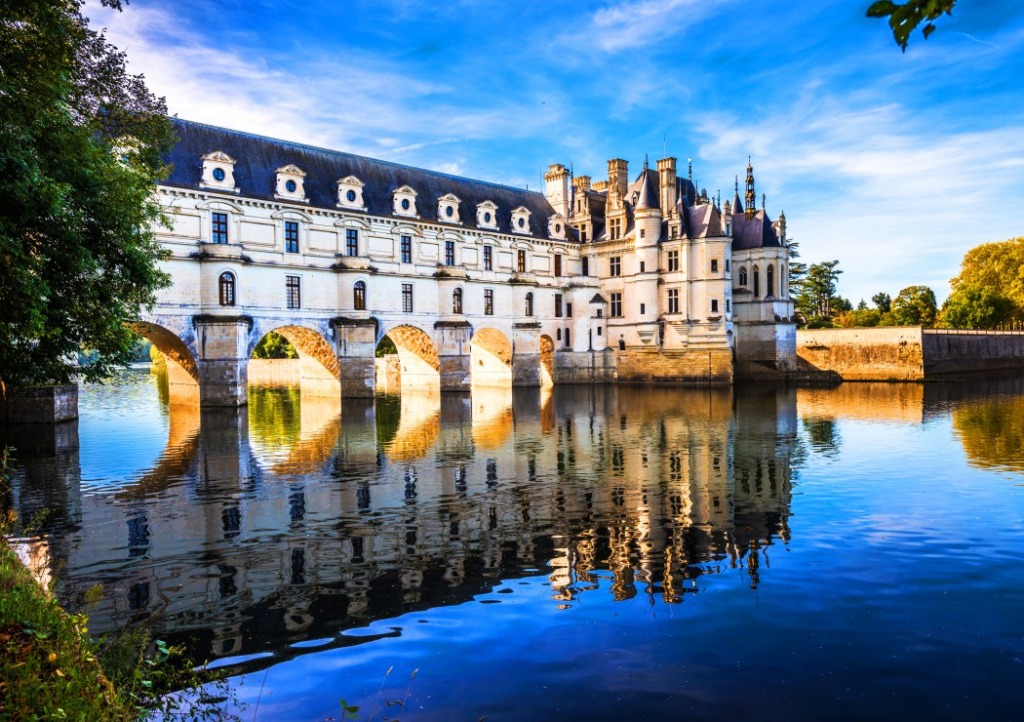 A low-angle view of the Chateau de Chenonceau spanning across a river under a vibrant blue sky.