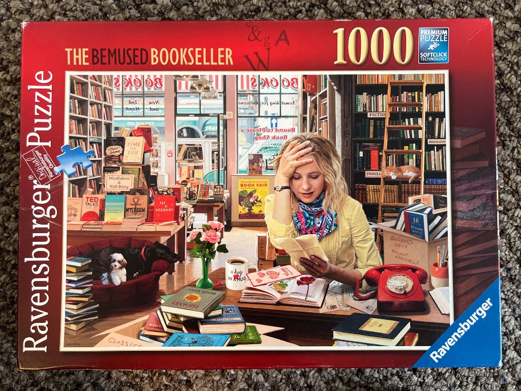 A bemused bookseller sits at a desk in a cluttered bookstore surrounded by books, a dog, and a cat.