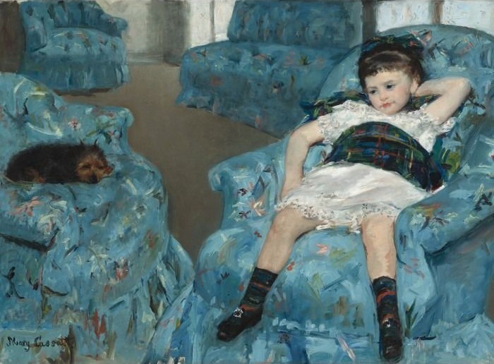 A young girl with dark hair lounging in a patterned blue armchair, with a small dog sleeping in an adjacent chair.