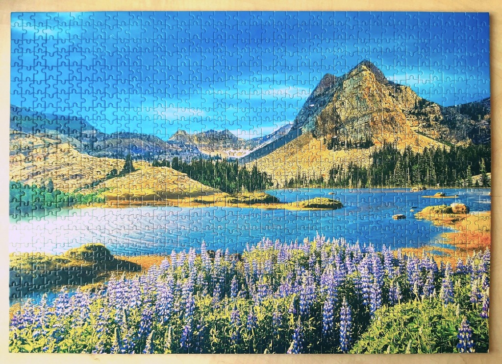 A scenic alpine landscape featuring a blue mountain lake surrounded by wildflowers and jagged peaks.