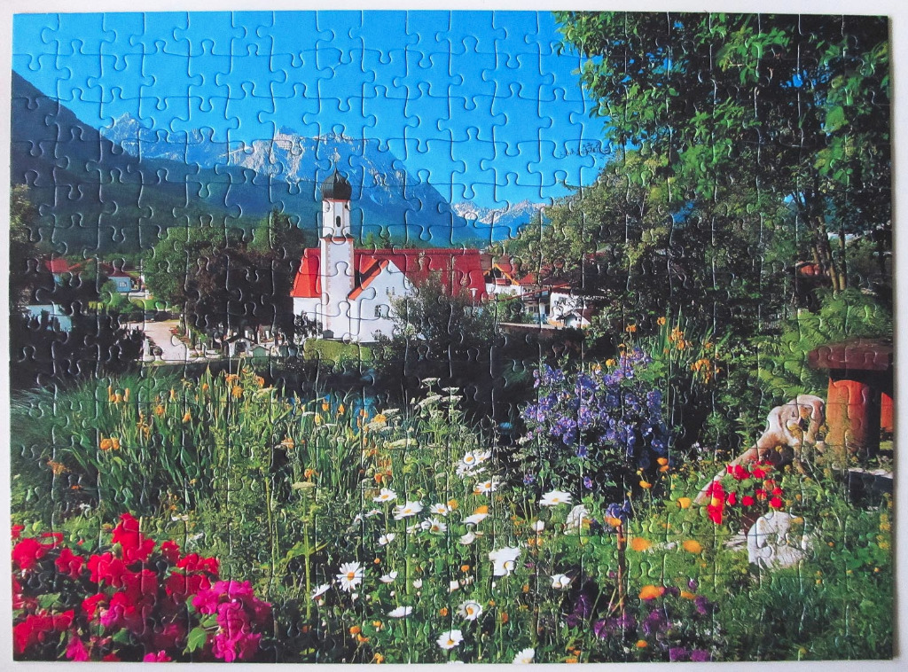 A sunny alpine landscape featuring a white church with red roofs and a vibrant flower garden in the foreground.