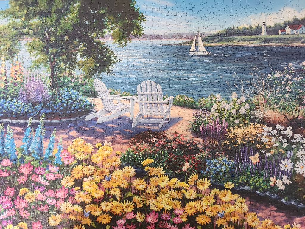 Two white Adirondack chairs overlooking a lake with a sailboat and lighthouse in a lush flower garden.