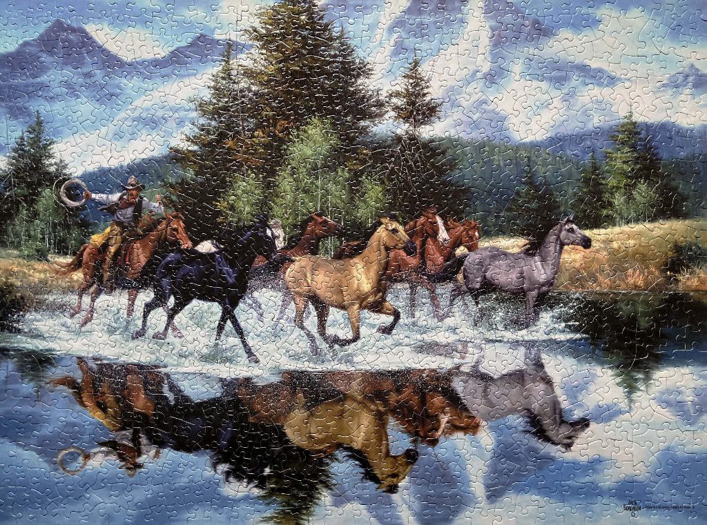 A cowboy on horseback and a herd of horses running through a shallow river with a mountain backdrop.