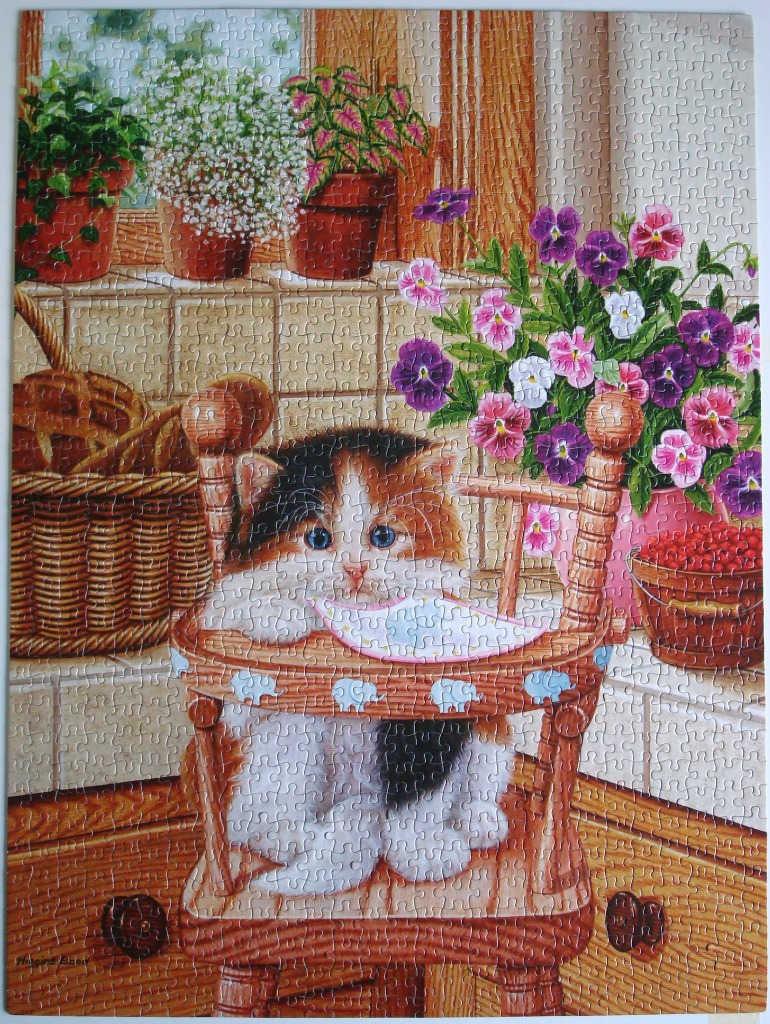 A fluffy calico kitten sits in a wooden high chair in a sunny kitchen filled with potted flowers and a basket of bread.