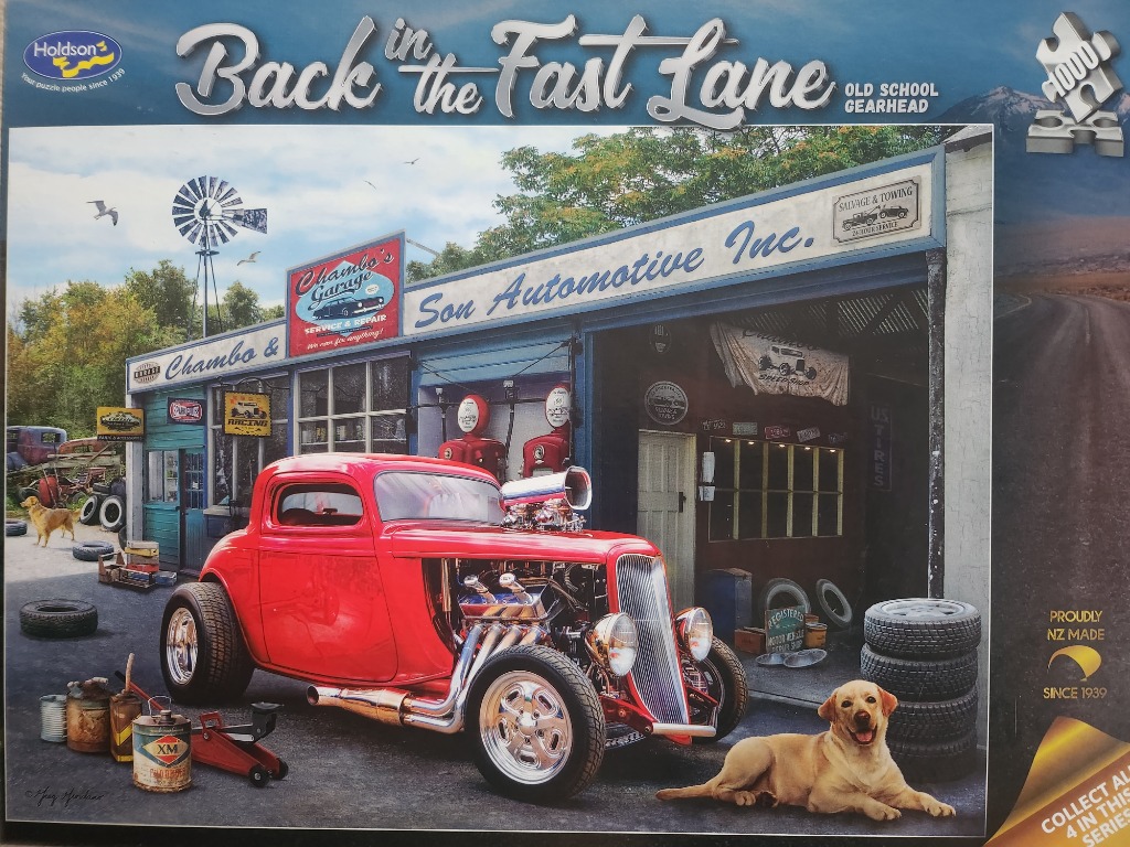 A red hot rod parked in front of a rustic automotive garage with a yellow dog resting on the pavement.