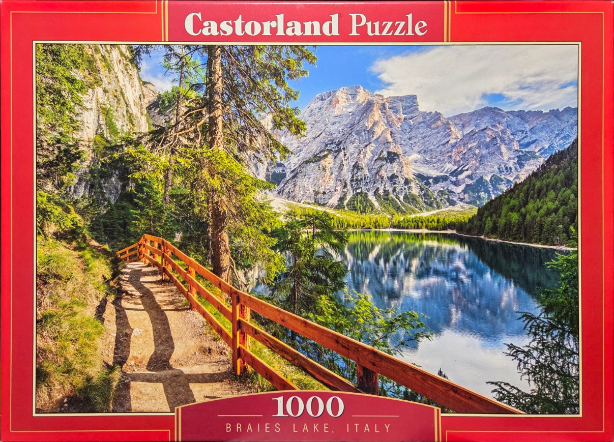 A scenic photograph of a wooden walkway along the edge of Braies Lake in Italy with high mountain peaks in the background.