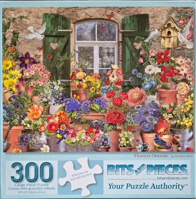 A collection of vibrant potted flowers arranged in front of a rustic stone cottage window with open green shutters.
