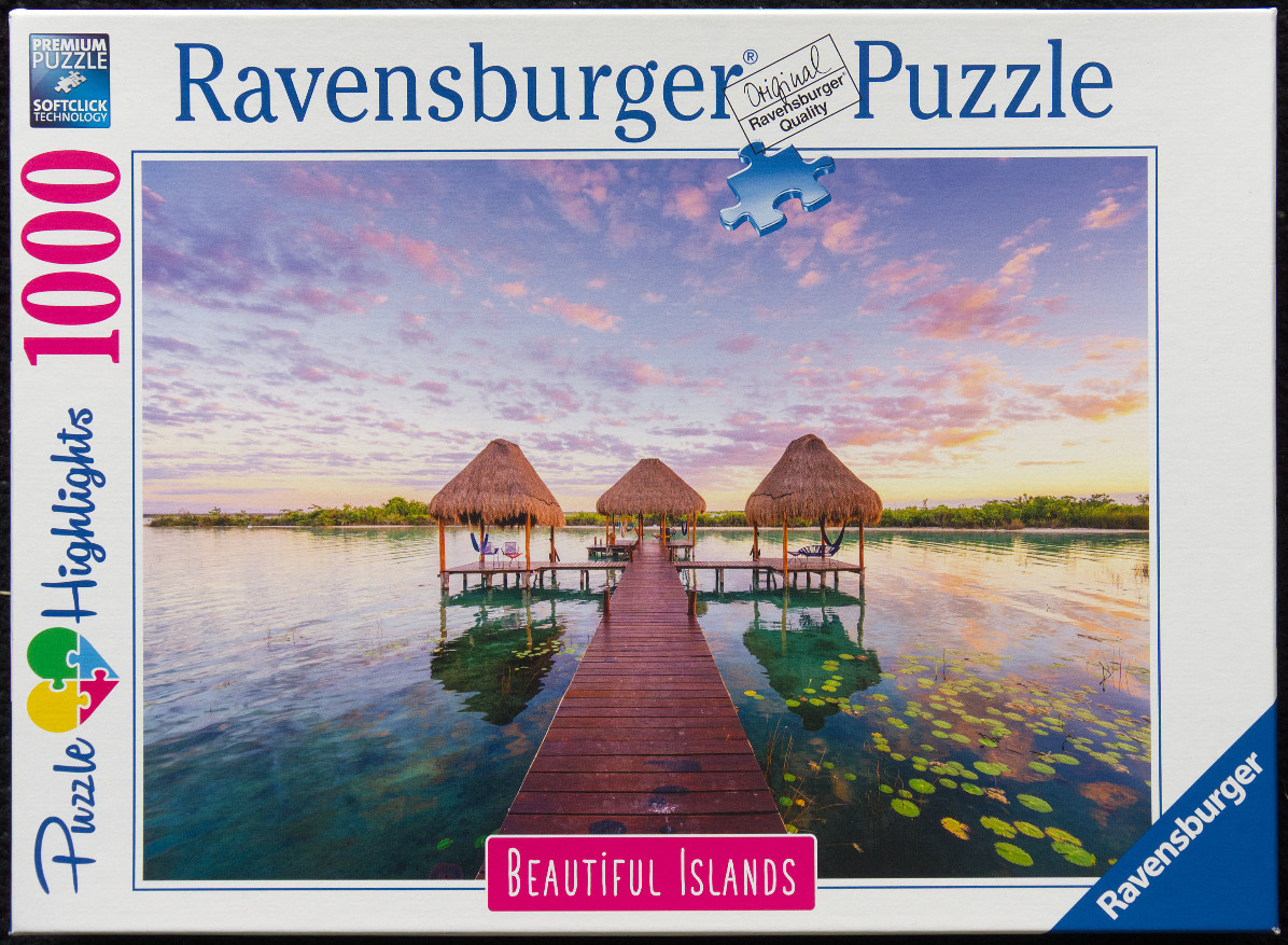 A wooden pier leading to three thatched-roof huts over a calm, clear tropical lagoon at sunset.